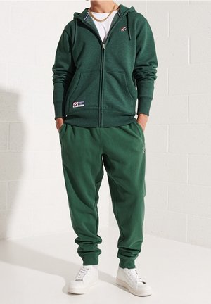 Felpa con zip verde in tessuto strutturato, con due tasche frontali e dettaglio logo. Pantaloni della tuta coordinati verdi con polsini elasticizzati. Scarpe da ginnastica bianche.