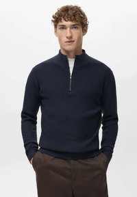 Marineblauer Reißverschluss-Pullover mit gerippter Struktur und Stehkragen. Kombiniert mit braunen Hosen, zeichnet er sich durch eine figurbetonte Silhouette und lange Ärmel aus.