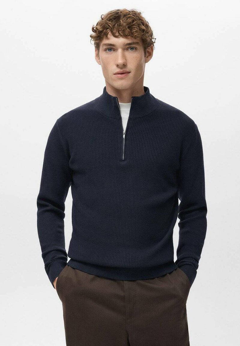 Marineblauer Reißverschluss-Pullover mit gerippter Struktur und Stehkragen. Kombiniert mit braunen Hosen, zeichnet er sich durch eine figurbetonte Silhouette und lange Ärmel aus.