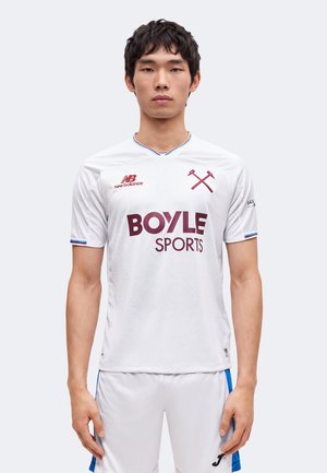 Jeune homme portant un maillot de football blanc avec le logo de Boyle Sports et un emblème de marteaux croisés, debout devant un fond clair uni.
