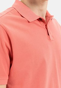 Koralfarvet polo shirt lavet af tekstureret bomuld. Har en klassisk krave, tre-knaps placket og korte ærmer.