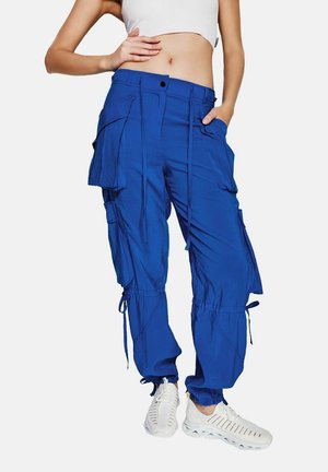 Femme portant un pantalon cargo bleu vif avec plusieurs poches et des chaussures de sport blanches, debout avec une main sur la hanche.
