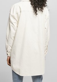 Chemise en velours côtelé beige avec col à bouton, manches longues et ourlet incurvé, présentant un tissu texturé et des détails de couture visibles.