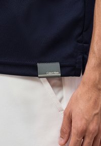 Gros plan d'une main posée sur le côté, portant un pantalon blanc et une chemise bleu marine avec une étiquette Calvin Klein visible.