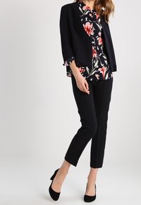 Blazer noir porté sur un chemisier à motifs floraux roses et blancs, accompagné d'un pantalon noir sur mesure et de talons noirs pointus.