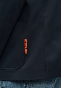 Τσέπη σκούρου ναυτικού μπλέ σακακιού με φωτεινή πορτοκαλί ετικέτα κειμένου «JUST CAVALLI», σε συνδυασμό με τζιν παντελόνι ανοιχτού μπλε χρώματος.