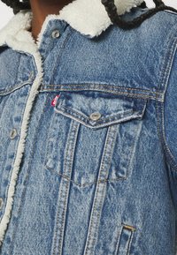 Veste en denim avec un col en polaire blanche douce, dotée de quincaillerie argentée, de deux poches poitrine et de coutures contrastantes en bleu.