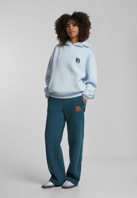 Felpa azzurra chiara con tasca frontale e logo blu navy, abbinata a pantaloni della tuta turchesi con logo arancione. Vestibilità morbida e rilassata.
