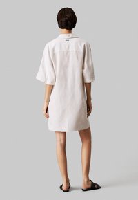 Witte linnen jurk met korte mouwen, relaxed fit en een notch kraag. Naaddetails en een klein logo accent aan de achterkant. Zwarte slide sandalen.