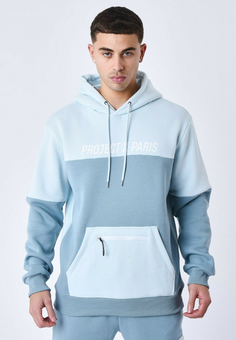 Sudadera azul claro con un bolsillo frontal, que presenta una sección con cremallera. Diseño de color bloque con tonos contrastantes de azul y texto en blanco.