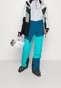 Lyžařský outfit obsahující vícebarevnou nepromokavou bundu a tyrkysové kalhoty s černými akcenty. Modelka drží bílou helmu.