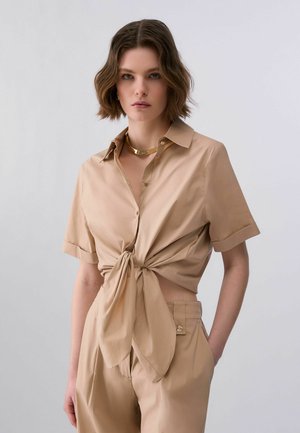 Kvinde med kort bølget hår iført beige kortærmet skjorte med knapper, bundet om taljen, og matchende højtaljede bukser mod lys baggrund.