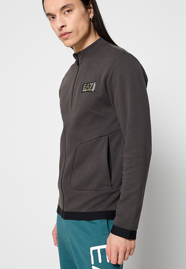 Giacca zip-up grigia scura con collo alto, due tasche frontali e un patch con logo sul petto. Realizzata in tessuto morbido e liscio.