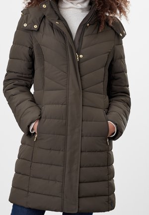Kvinde iført en lang, mørke olivengrøn quiltet vinterjakke med lynlås og trykknapper, hænderne i lynlåslommer, kombineret med en lysegrå rullekrave.
