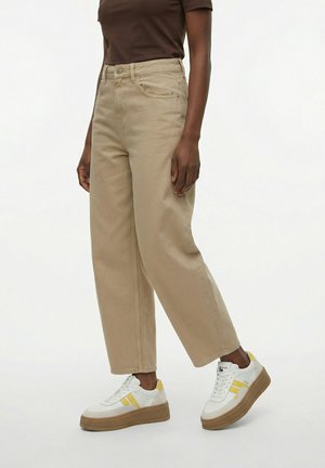 Persona vistiendo pantalones anchos beige, zapatillas blancas con rayas amarillas y una camiseta marrón de manga corta, de pie contra un fondo liso.