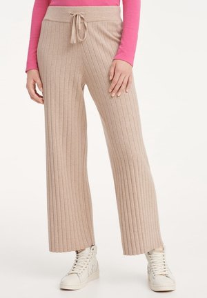 Frau trägt beige gerippte, weite Hosen mit Kordelzug in der Taille und weißen High-Top-Sneakers, kombiniert mit einem rosa Langarmshirt.