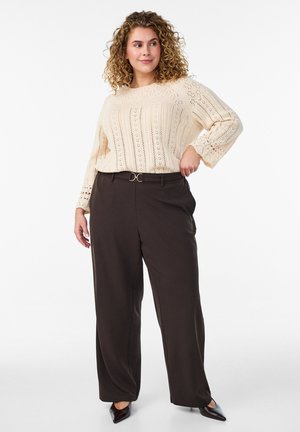 Žena s kučeravými vlasmi, oblečená v krémovom pletenom svetri a tmavých širokých nohaviciach s vysokým pásom a opaskom, pózujúca s rukou na bokoch, pozriaca sa bokom.