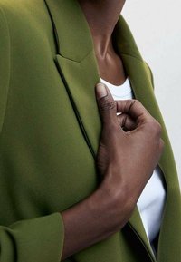 Blazer vert à la coupe ajustée, doté d'un revers acéré et d'une texture lisse. Chemise blanche en dessous, main ajustant le blazer.