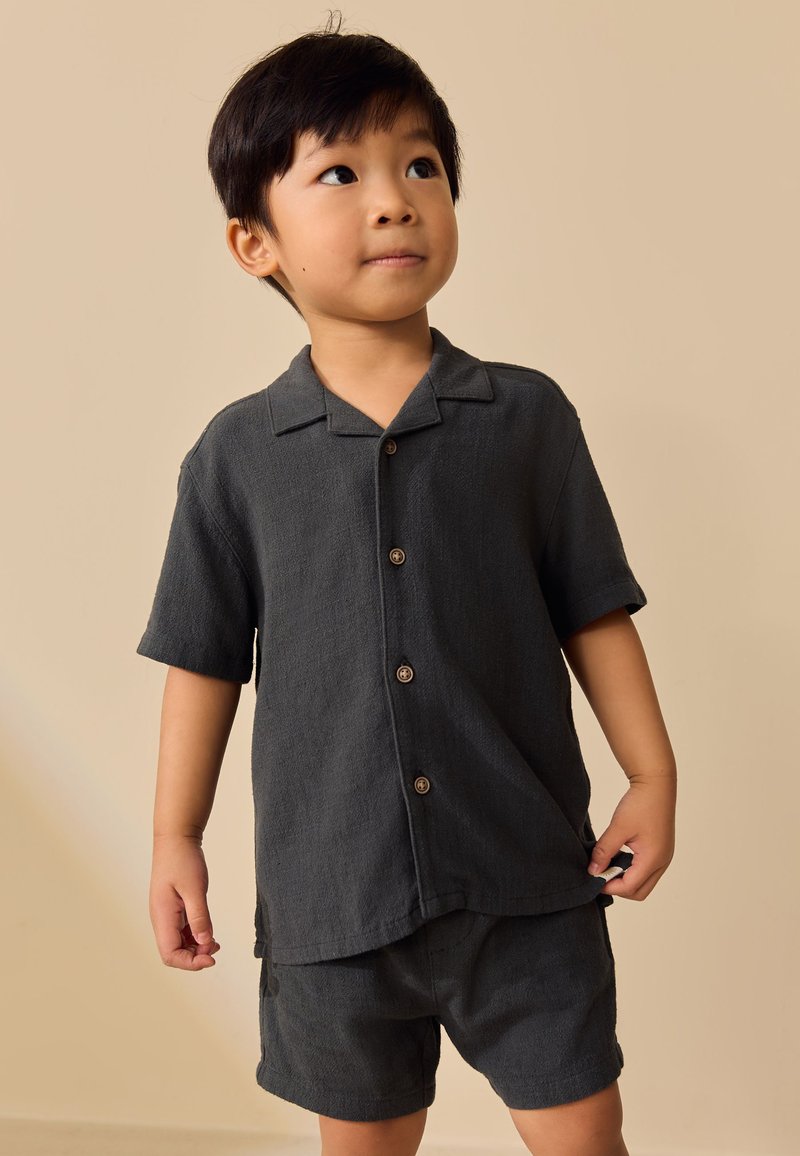 Niño pequeño con camisa de manga corta abotonada de color gris oscuro y pantalones cortos a juego, de pie sobre un fondo beige neutro.