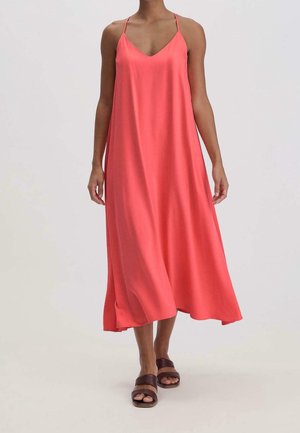 Kobieta w koralowo-różowej, bezrękawnej sukience midi z cienkimi ramiączkami i brązowych płaskich sandałach, stojąca na neutralnym tle.