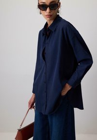 Camicia blu navy con bottoni, vestibilità comoda, maniche lunghe e tasca. Abbinata a pantaloni in denim scuro e una borsa marrone.