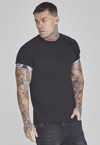 Melna t-kreklu ar izspiestu "SIKSILK" logo, īsām raglan piedurknēm ar ziedu rakstu un pieguļošu dizainu, valkā tetovēts vīriešu modelis.