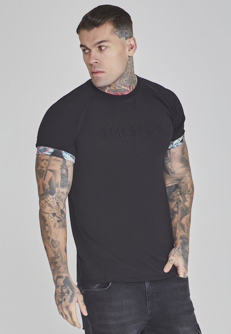 Melna t-kreklu ar izspiestu "SIKSILK" logo, īsām raglan piedurknēm ar ziedu rakstu un pieguļošu dizainu, valkā tetovēts vīriešu modelis.