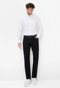Camicia bianca a bottone con taschino, abbinata a pantaloni neri e una cintura nera. Il modello indossa sneakers bianche. Design semplice e pulito.