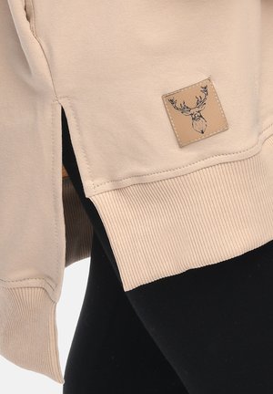 Beige sweatshirt hem with ribbed cuff and side slit, featuring a small stitched patch with a black outlined deer head.