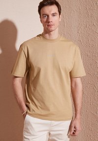 Tan bomulds-T-shirt med korte ærmer, rund hals og broderet logo på brystet; modellen bærer lyse bukser mod en tekstureret baggrund.