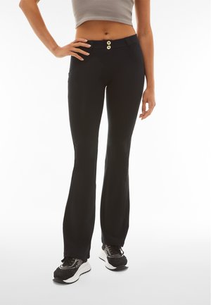 Nainen, joka pukee mustat liehuvat housut, joissa on kaksinkertainen napitus, harmaa lyhyt paita ja mustat lenkkarit, seisoo käsi toisella lantiolla.