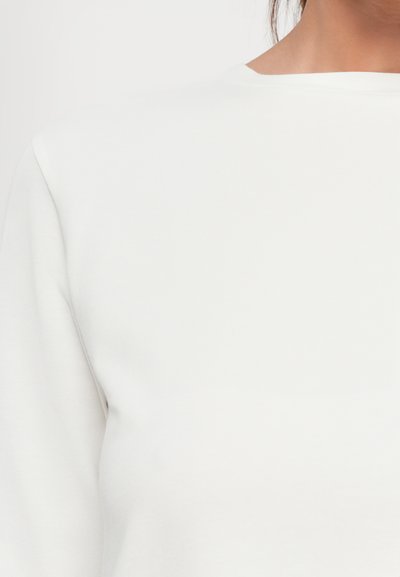 Close-up of a person wearing a plain white crew-neck long-sleeve shirt against a neutral background.