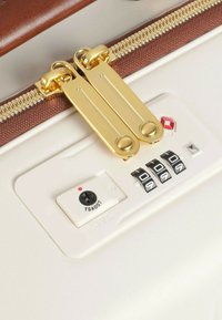 Valigia bianca con cerniera dorata, dettagli marroni e serratura combinata. La visualizzazione include un lucchetto TSA e dischi numerici per impostare il codice.