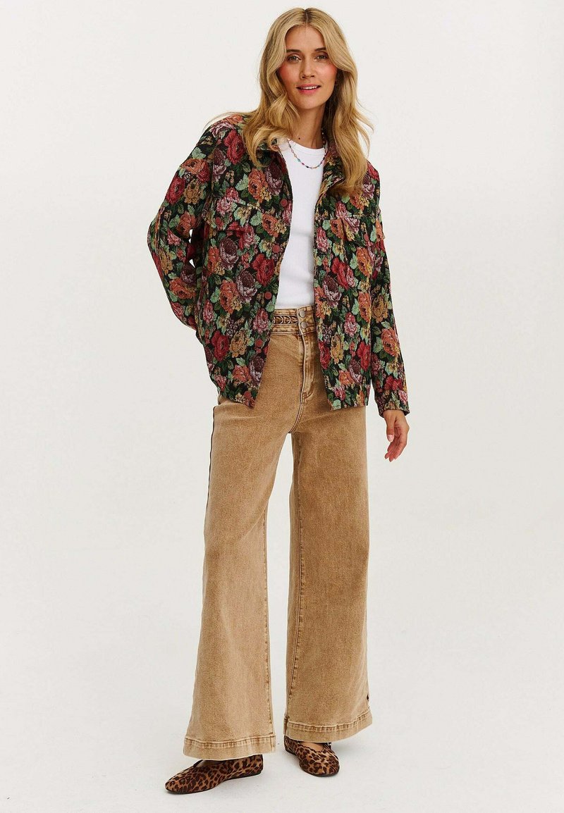 Kurtka w kwiatowy wzór w kolorach zielonym, kremowym i czerwonym, zestawiona z beżowymi spodniami o wysokim stanie z szerokimi nogawkami oraz brązowymi butami w panterkę.