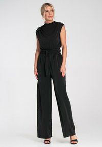 Czarny kombinezon z krótkimi rękawami i wysokim dekoltem. Posiada elastyczny pas z tkaninowym paskiem. Szerokie nogawki, gładka tekstura.