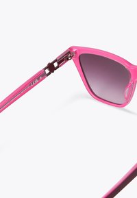 Occhiali da sole rosa con montatura triangolare, lenti grigie sfumate e dettagli trasparenti all'articolazione. Le aste presentano un elemento di design unico.