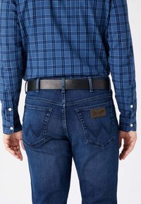 Donkerblauwe denim jeans met een leren Wrangler-label; gedragen met een blauwe geruite overhemd met knopen. Zwarte riem zichtbaar op de taille.