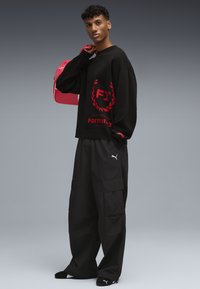 Felpa nera con logo F1 rosso e design di alloro, abbinata a pantaloni cargo neri larghi. Il modello tiene una borsa rosa con dettagli bianchi.