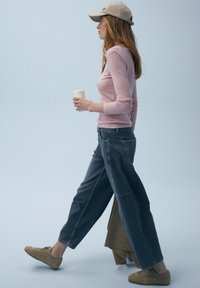 Haut à manches longues rose clair, jeans larges bleu foncé, baskets beiges, portant une casquette beige, tenant une tasse de café, marchant devant un fond bleu doux.