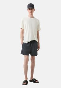 Lys beige kortærmet t-shirt; mørkegrå badebukser med snøre; sorte slide sandaler; tætsiddende kasket. Enkel, afslappet outfit.