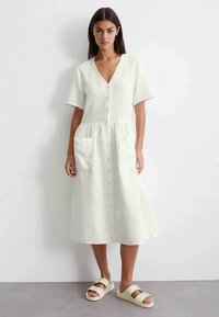 Frau, die an einer weißen Wand steht, trägt ein weißes Midi-Kleid mit Knöpfen und kurzen Ärmeln sowie zwei vorderen Taschen, und beige Sandalen.