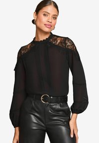 Blusa nera con dettagli in pizzo sul collo e sulle spalle, caratterizzata da accenti plissettati con bottoni. Abbinata a pantaloni in pelle nera a vita alta.