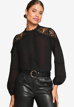 Blusa nera con dettagli in pizzo sul collo e sulle spalle, caratterizzata da accenti plissettati con bottoni. Abbinata a pantaloni in pelle nera a vita alta.