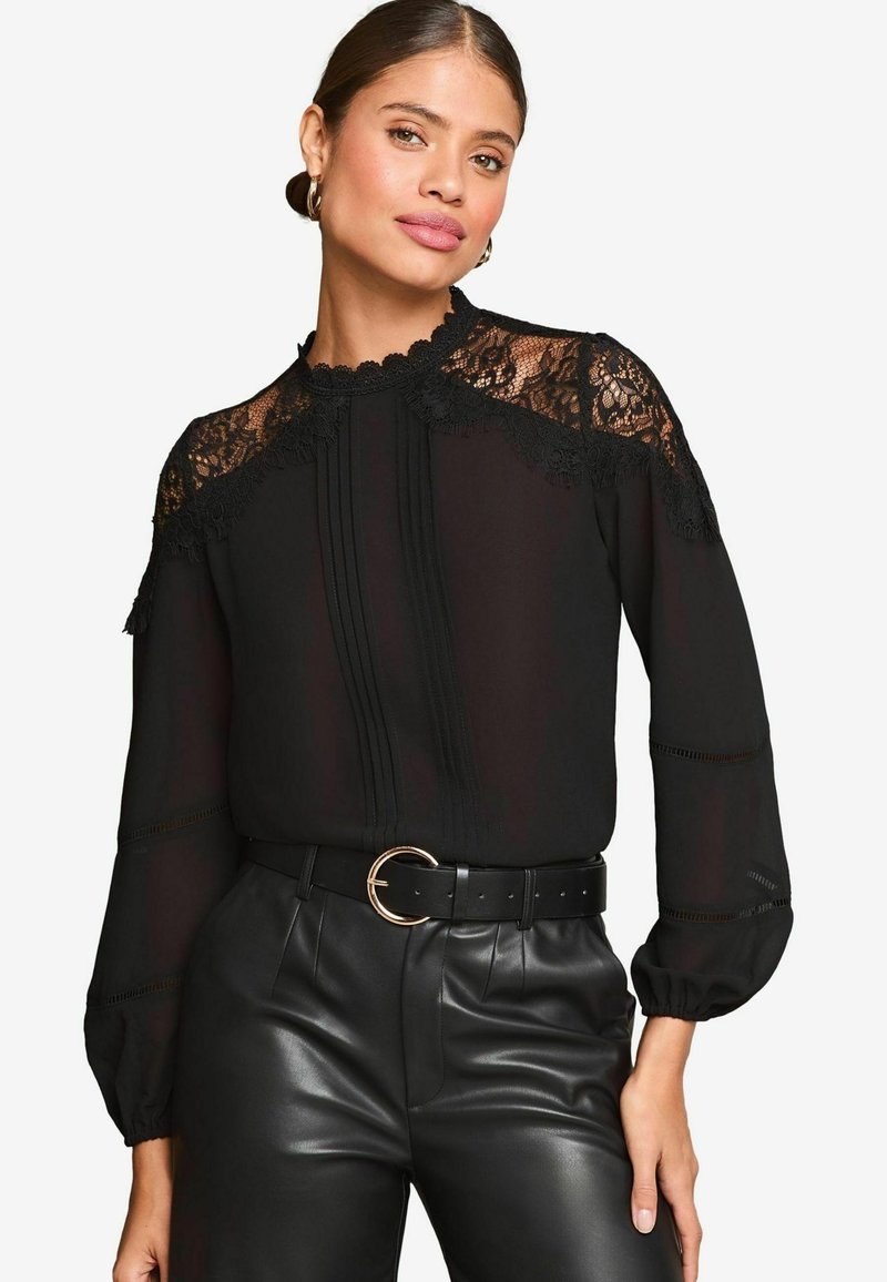 Blusa nera con dettagli in pizzo sul collo e sulle spalle, caratterizzata da accenti plissettati con bottoni. Abbinata a pantaloni in pelle nera a vita alta.