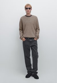 Brown textured sweater, black jeans, and black sneakers. Model wears black sunglasses; hands in pockets against a plain background.
