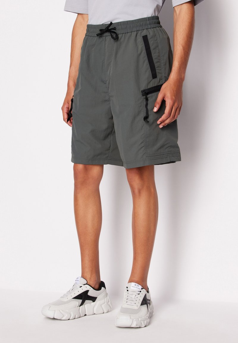 Pantaloni corti grigi leggeri con vita elastica, tasche con zip nere e vestibilità rilassata. Abbinati a scarpe da ginnastica grigie e nere.