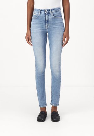 Person iført lyseblå skinny jeans med høj talje og sorte slip-on sko, stående mod en ensfarvet hvid baggrund.