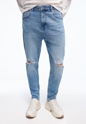 Light blue ripped skinny jeans worn with white chunky sneakers and a beige sweater, shown from waist to feet on a white background.