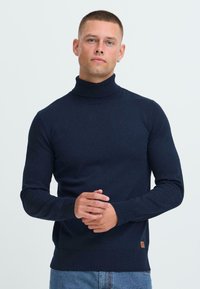 Joven con el cabello corto, vistiendo un suéter de cuello alto azul marino y jeans de mezclilla claros, de pie contra un fondo liso claro.