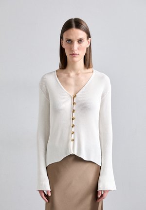 Cardigan en mailles blanches à côtes, avec un col en V, comportant six boutons dorés sur le devant et des manches évasées. Texture lisse et ourlet cropped.